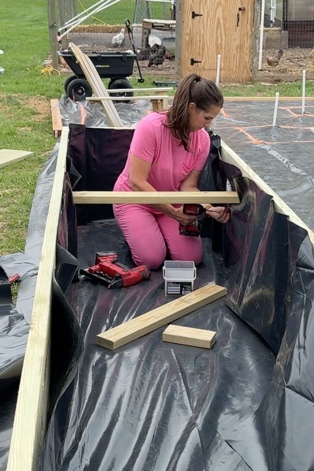 A woman in a pink outfit kneels inside a black-lined raised garden bed frame and drills an interior support brace into place, with a nail gun, screws, and extra lumber pieces nearby and free-range chickens and a garden cart visible in the background.