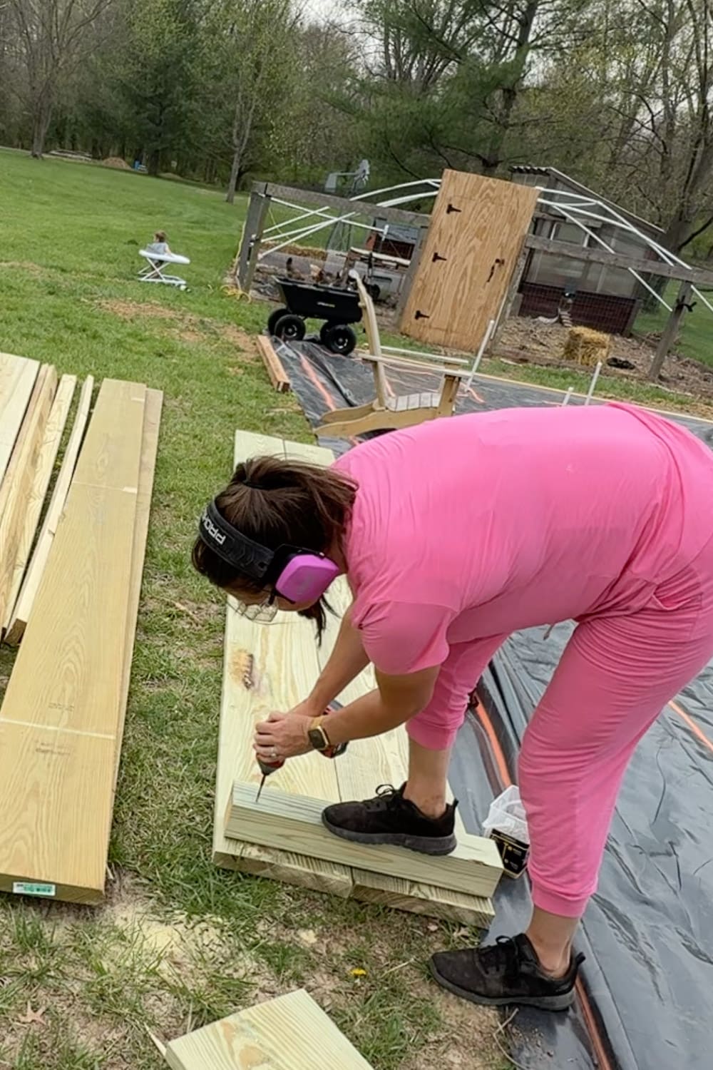 A woman in a pink outfit and pink ear protection uses a power drill to assemble pressure-treated lumber boards, with a chicken coop, garden cart, and black weed barrier visible in the background.