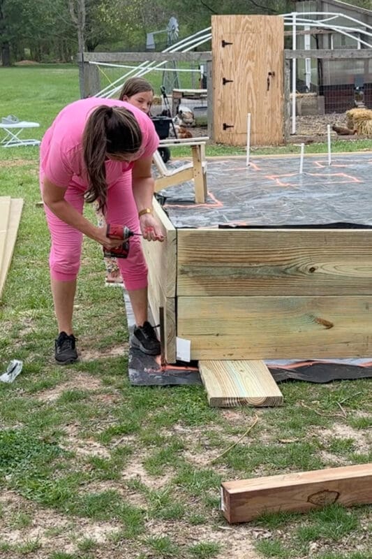 A woman in a pink outfit uses a power drill to fasten boards on a partially constructed raised garden bed, while a girl assists from behind. The weed barrier layout with orange spray paint markings and a chicken coop are visible in the background.