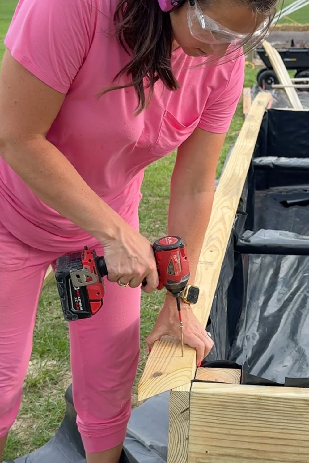 A close-up of a woman in a pink outfit and safety glasses using a Milwaukee impact driver to drive a screw into the top cap board of a pressure-treated raised garden bed, with the black liner and garden cart visible in the background.