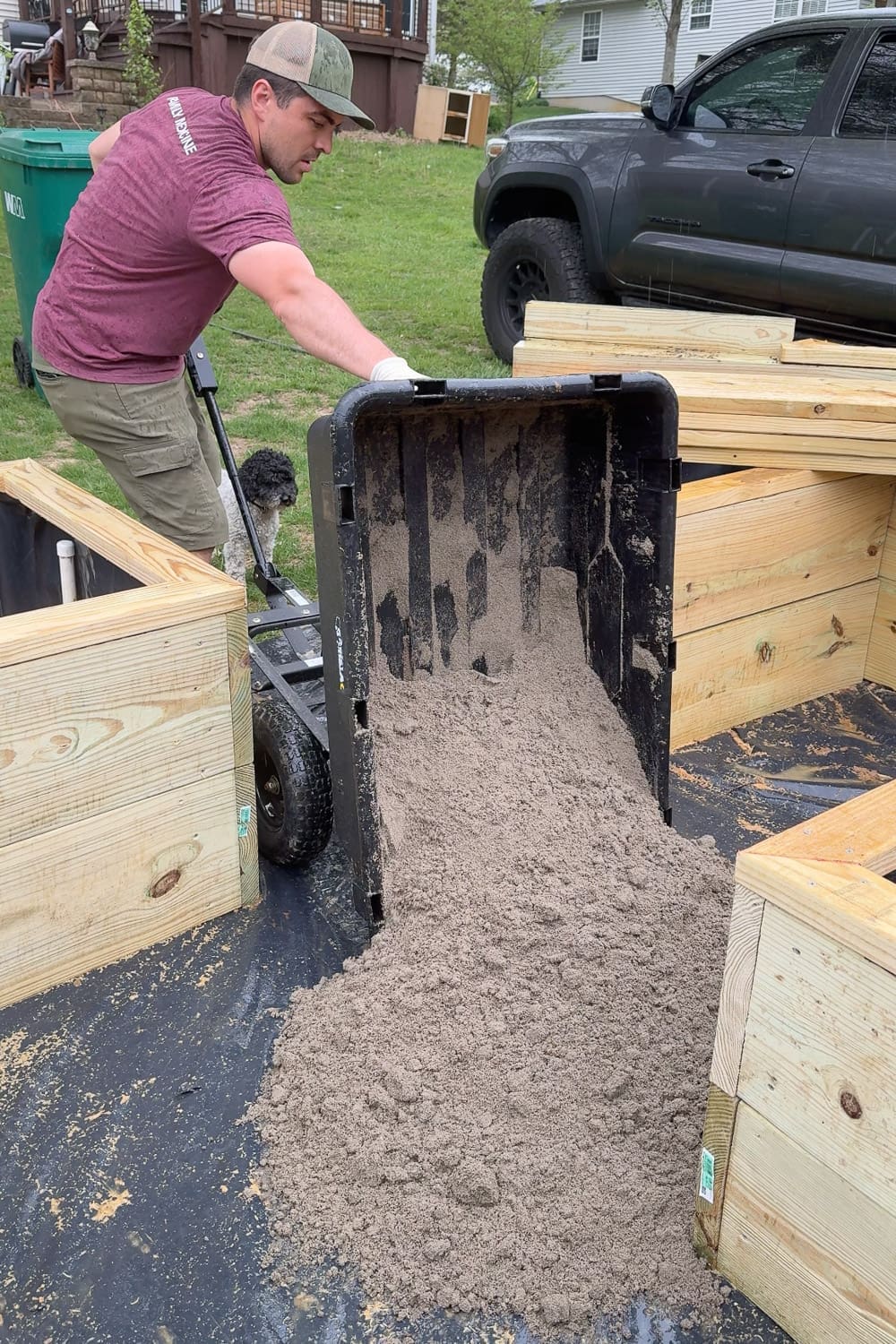 Dumping paver base sand from a utility cart onto landscape fabric between raised garden boxes to prep the walkway area