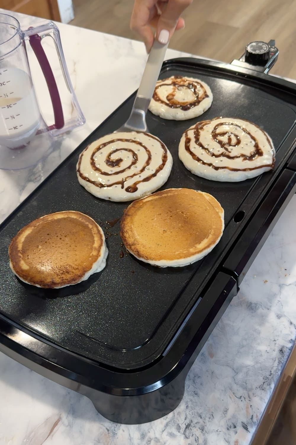 Hand using a metal spatula to flip cinnamon roll pancakes on a black electric griddle, showing two already-flipped golden brown pancakes alongside two unflipped pancakes with visible cinnamon swirl patterns on top.