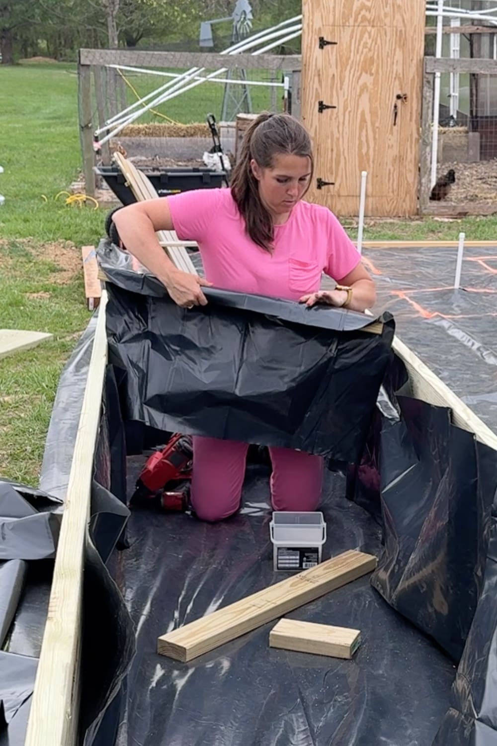 A woman in a pink outfit kneels inside a large raised garden bed frame and manually folds and positions the black liner fabric against the interior walls, with a nail gun, screws, and lumber pieces on the liner floor and a chicken coop visible in the background.