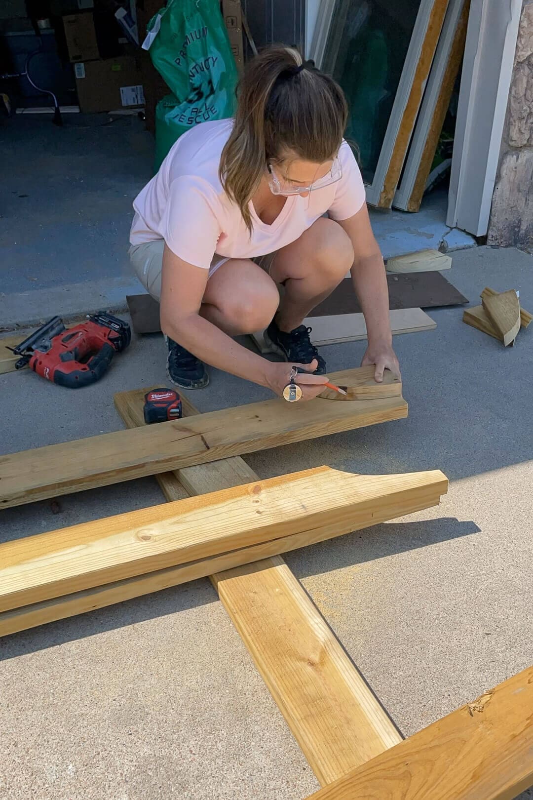 A woman wearing a pink shirt and black sneakers crouches on a concrete driveway, measuring and marking a wooden beam with a red tape measure. The project is laid out with multiple wooden planks, forming the base structure of a woodworking project. In the background, an open garage with tools, supplies, and a child’s bicycle is visible.