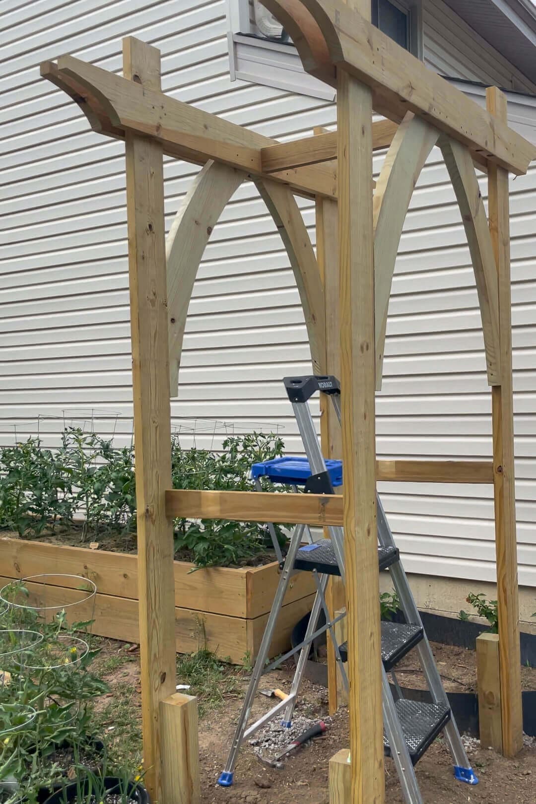 A nearly completed wooden garden arbor with decorative curved beams and a sturdy frame. A step ladder and various tools lay around the base, indicating final construction touches.