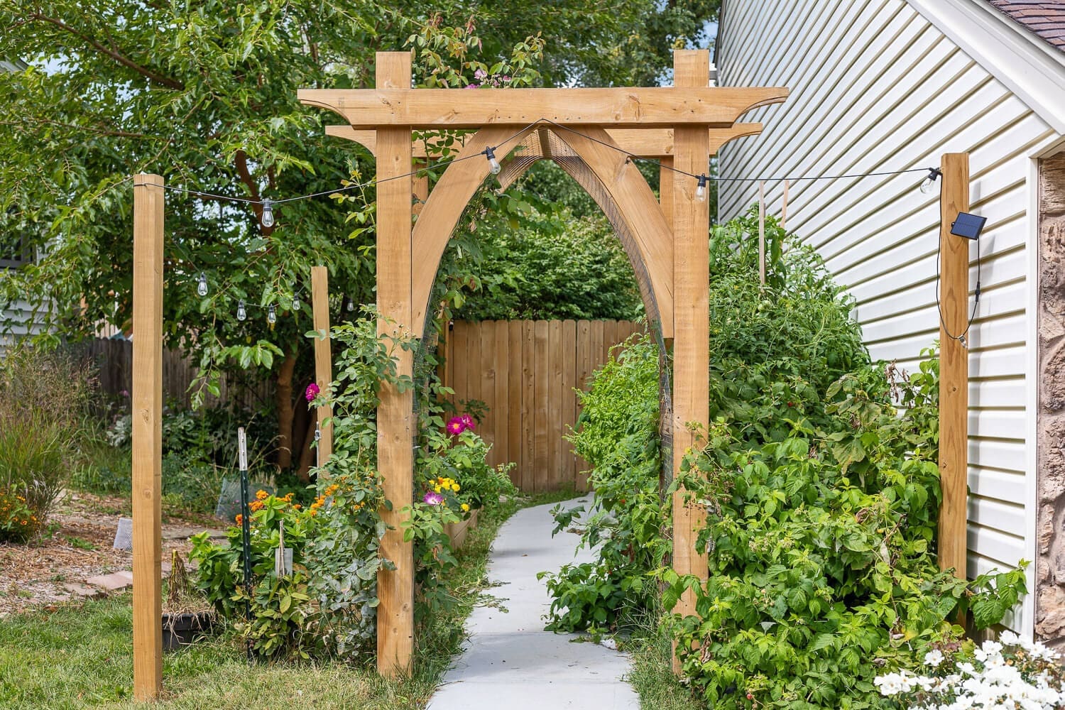 A wooden garden arbor with elegant arched detailing, covered in climbing vines with pink flowers. The arbor stands at the entrance to a lush backyard garden, surrounded by raised beds and string lights.