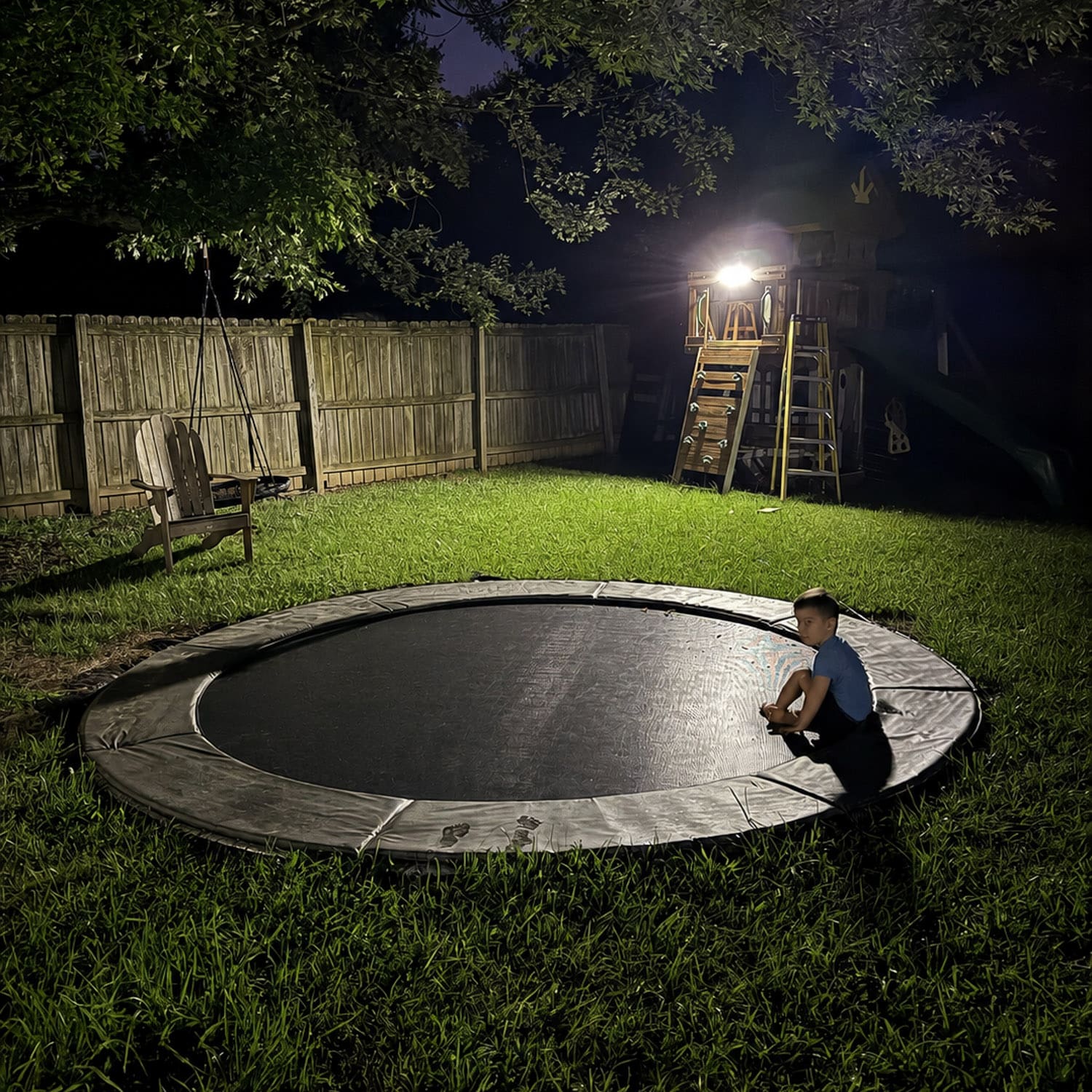 Finished in-ground trampoline sunk flush with the backyard grass at night, a boy resting on the padded rim, showing the result of learning how to bury a trampoline.