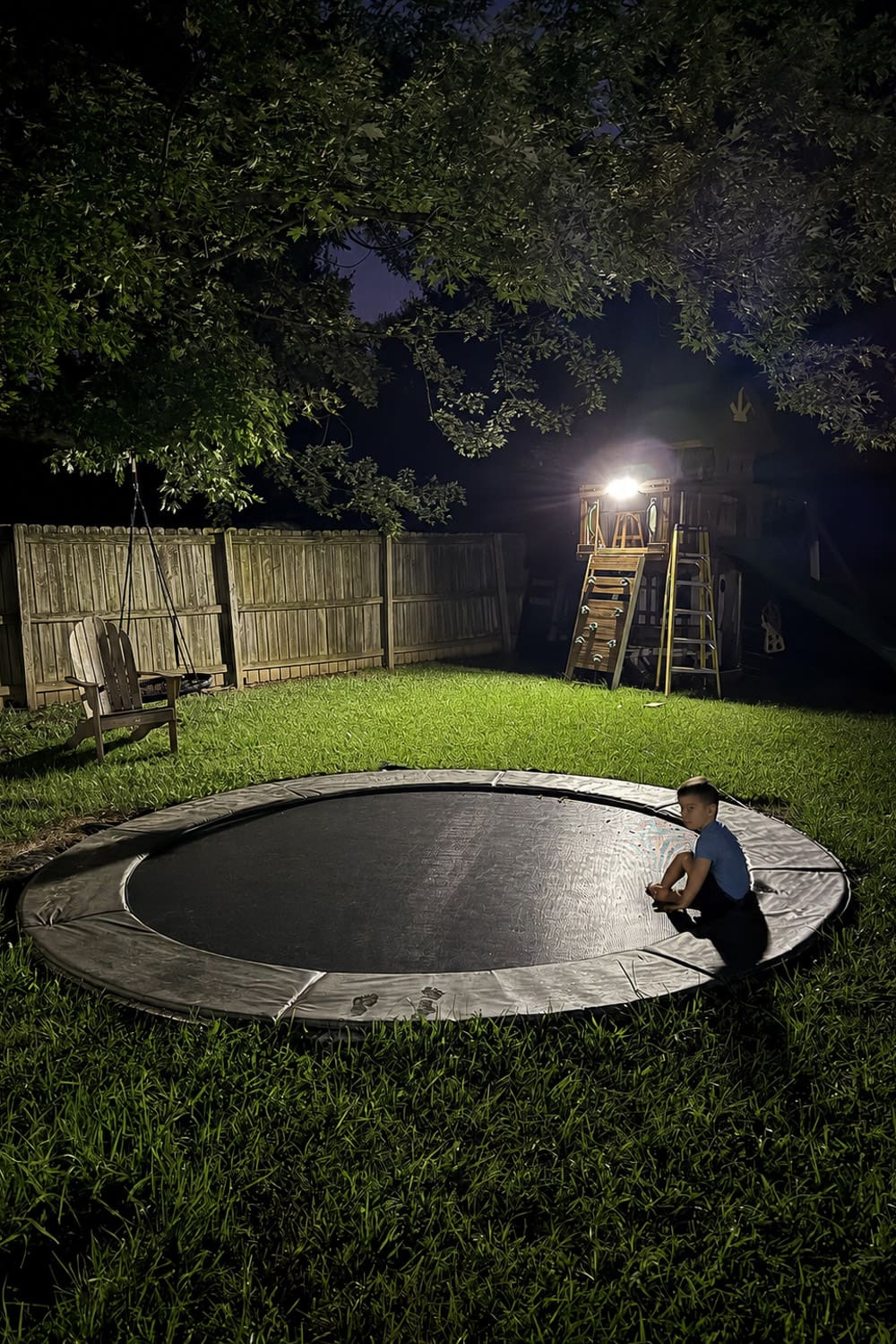 Buried trampoline sitting level with the lawn after dark, a boy on the padded edge near a lit playset, the completed result of how to bury a trampoline.