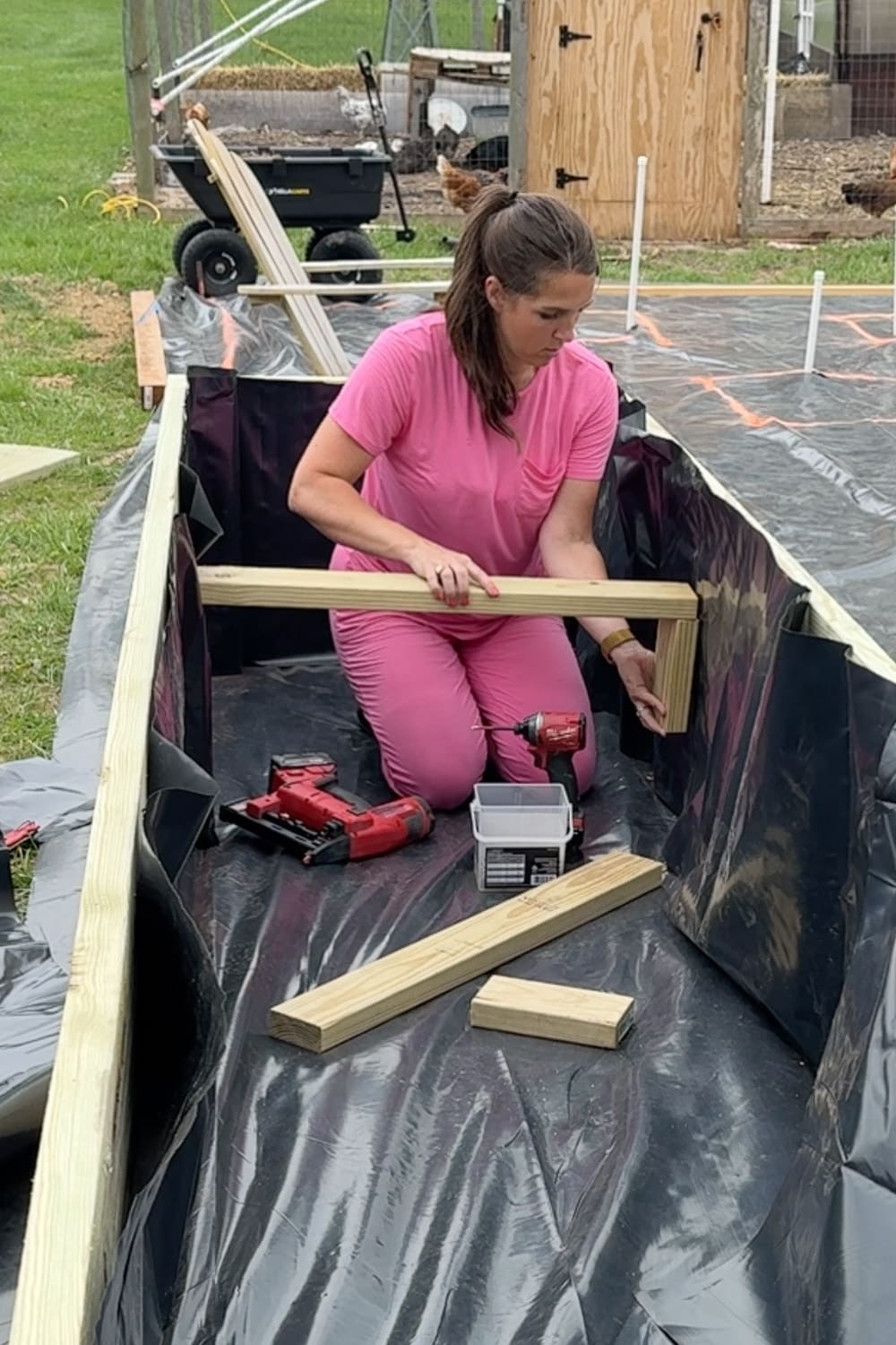A woman in a pink outfit kneels inside a large raised garden bed frame lined with black fabric, using a red impact driver to install an interior cross-support brace, with a nail gun and box of screws on the liner nearby and a chicken coop visible in the background.