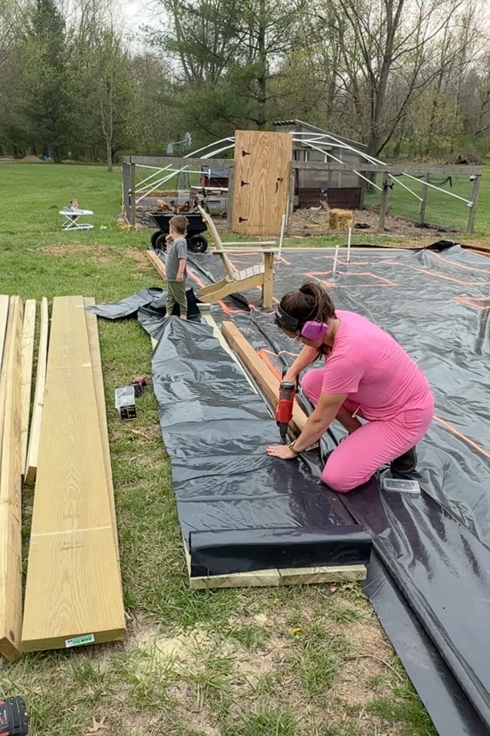 A woman in a pink outfit and pink ear protection kneels on black weed barrier fabric to drill the first board of a tall planter box into place, while a young boy stands nearby on the layout area watching. Stacked lumber, a chicken coop, and a cart are visible in the background.