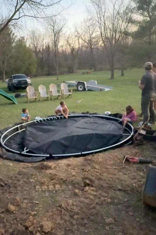 Attaching the trampoline mat and springs while installing an in-ground trampoline with family helping in the backyard