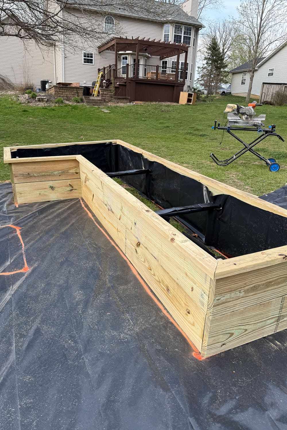 An L-shaped raised garden bed built from pressure-treated lumber and lined with black fabric, positioned on weed barrier in a backyard. A house with a pergola-covered deck and a miter saw on a stand are visible in the background.