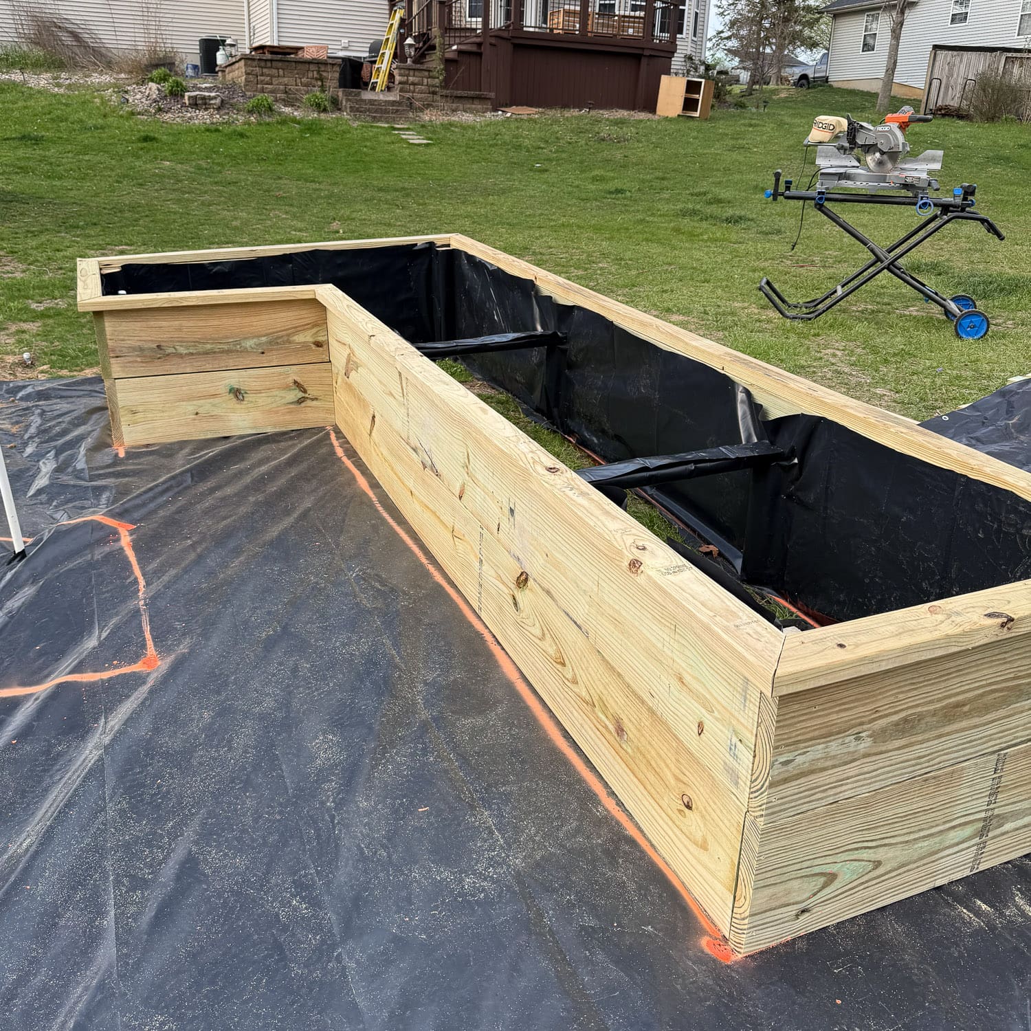 Two partially constructed L-shaped raised garden beds made from pressure-treated lumber, lined with black landscape fabric, sitting on black weed barrier in a backyard. A miter saw on a stand is visible in the background near a house with a deck.