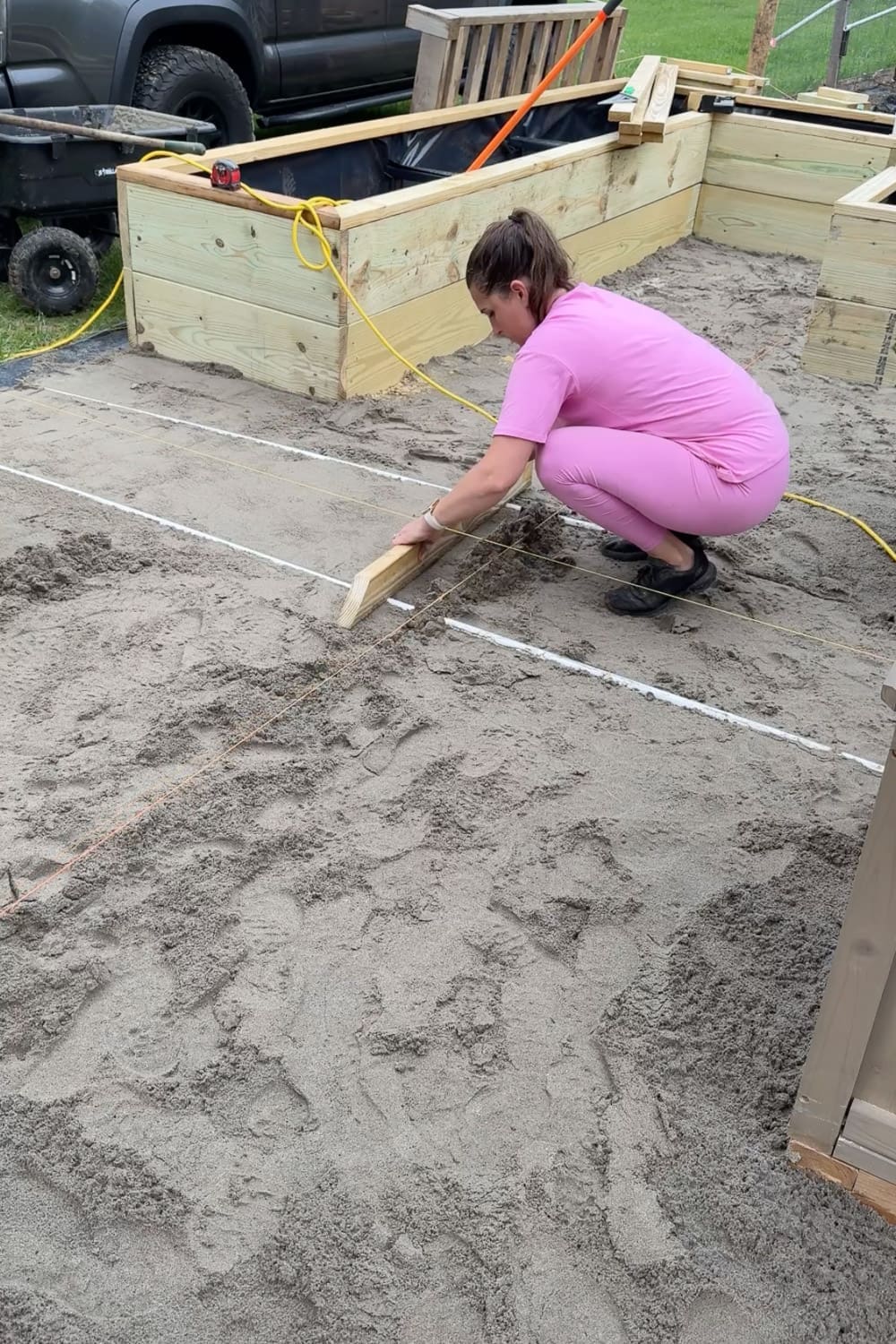Using a wooden board to mark straight grid lines in the sand base, creating a guide for laying square pavers in a checkerboard pattern