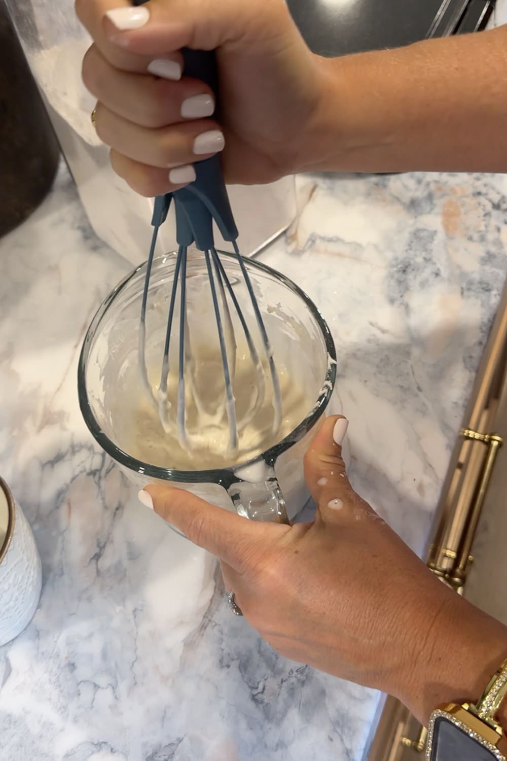 Hands whisking pancake batter in a glass measuring cup with a blue silicone whisk on a marble countertop, mixing the batter for cinnamon roll pancakes made with pancake mix.