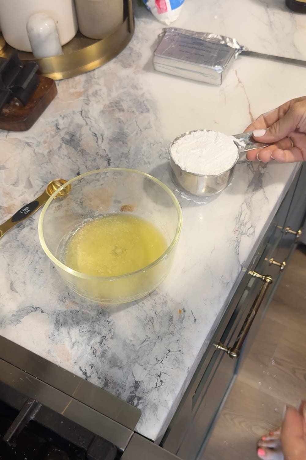 Hand holding a metal measuring cup heaped with powdered sugar next to a glass bowl of melted butter on a marble countertop, preparing the ingredients for homemade cream cheese glaze.