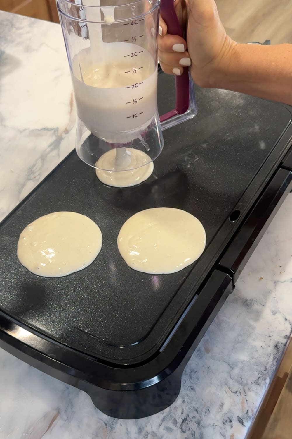 Hand using a pancake batter dispenser to pour two round circles of pancake batter onto a black electric griddle, the first step in cooking cinnamon roll pancakes.