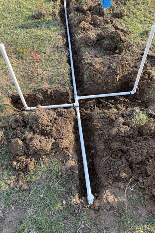 Close-up of a PVC cross fitting branching out to multiple spigot risers in the trench for a DIY raised bed irrigation system