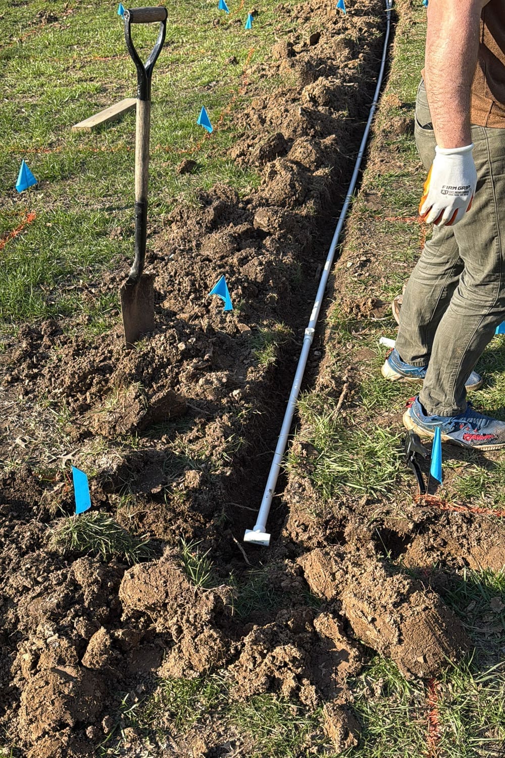 PVC pipe and tee fittings laid out in a freshly dug trench, ready to be buried as part of a raised bed garden irrigation system