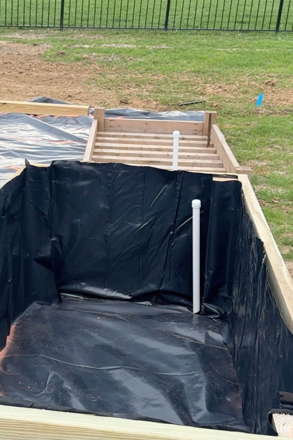Wooden raised garden bed lined with black landscape fabric showing two PVC watering spigots positioned inside the box, ready for soil