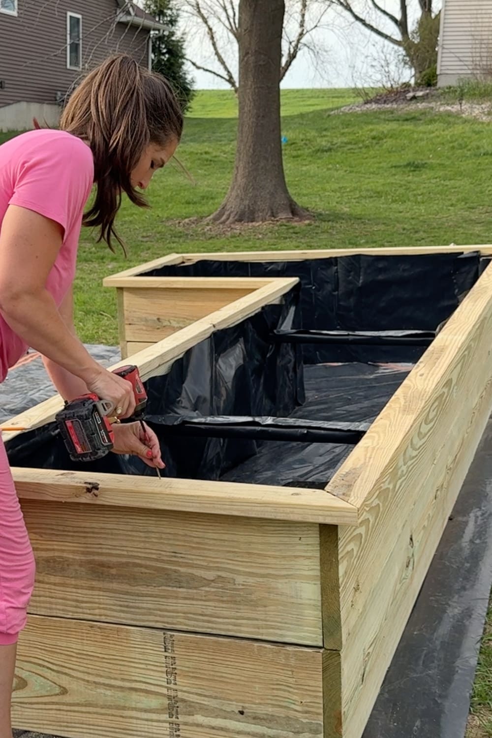 A woman in a pink outfit uses a Milwaukee impact driver to secure the black liner fabric under the top cap board of a nearly finished L-shaped raised garden bed made from pressure-treated lumber, with a second smaller bed and a grassy backyard visible in the background.
