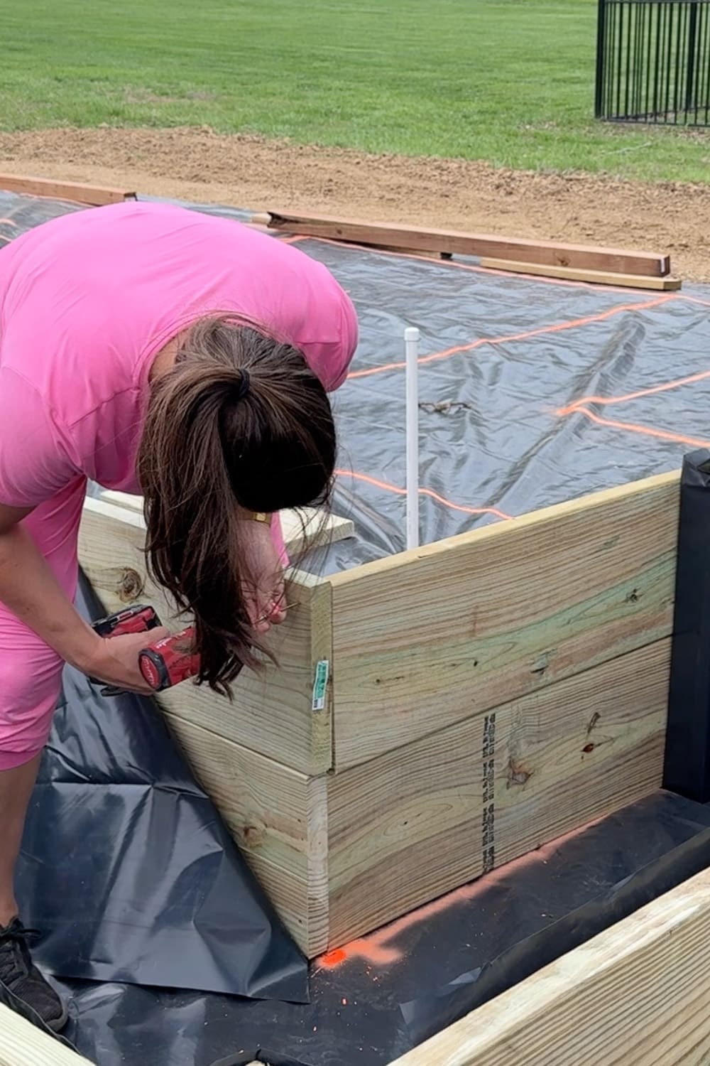 A woman in a pink outfit leans over the corner of a nearly completed raised garden bed, using a red impact driver to fasten the final boards of the corner joint, with the weed barrier layout and a white PVC stake visible in the background.