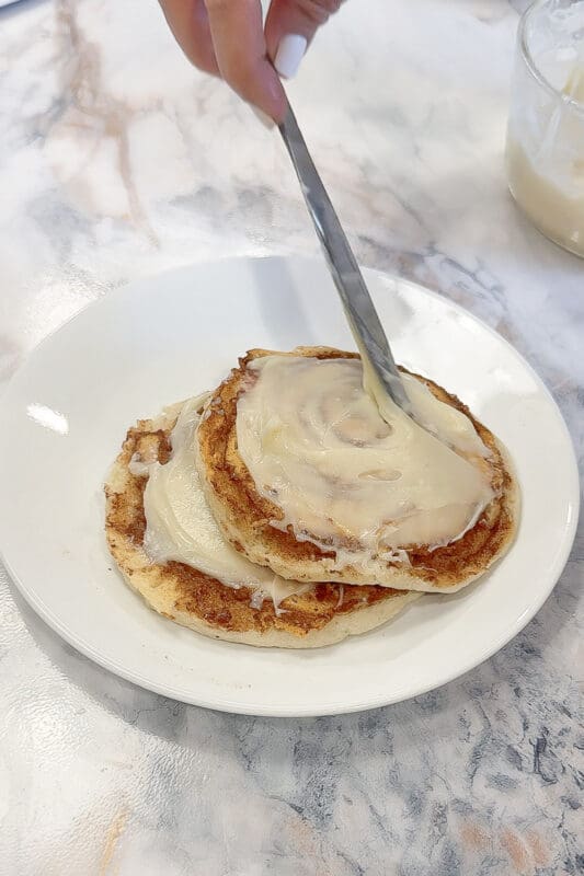 Hand spreading thick homemade cream cheese glaze over a stack of two cinnamon roll pancakes on a white plate using a butter knife, with a bowl of extra glaze visible in the background on a marble countertop.