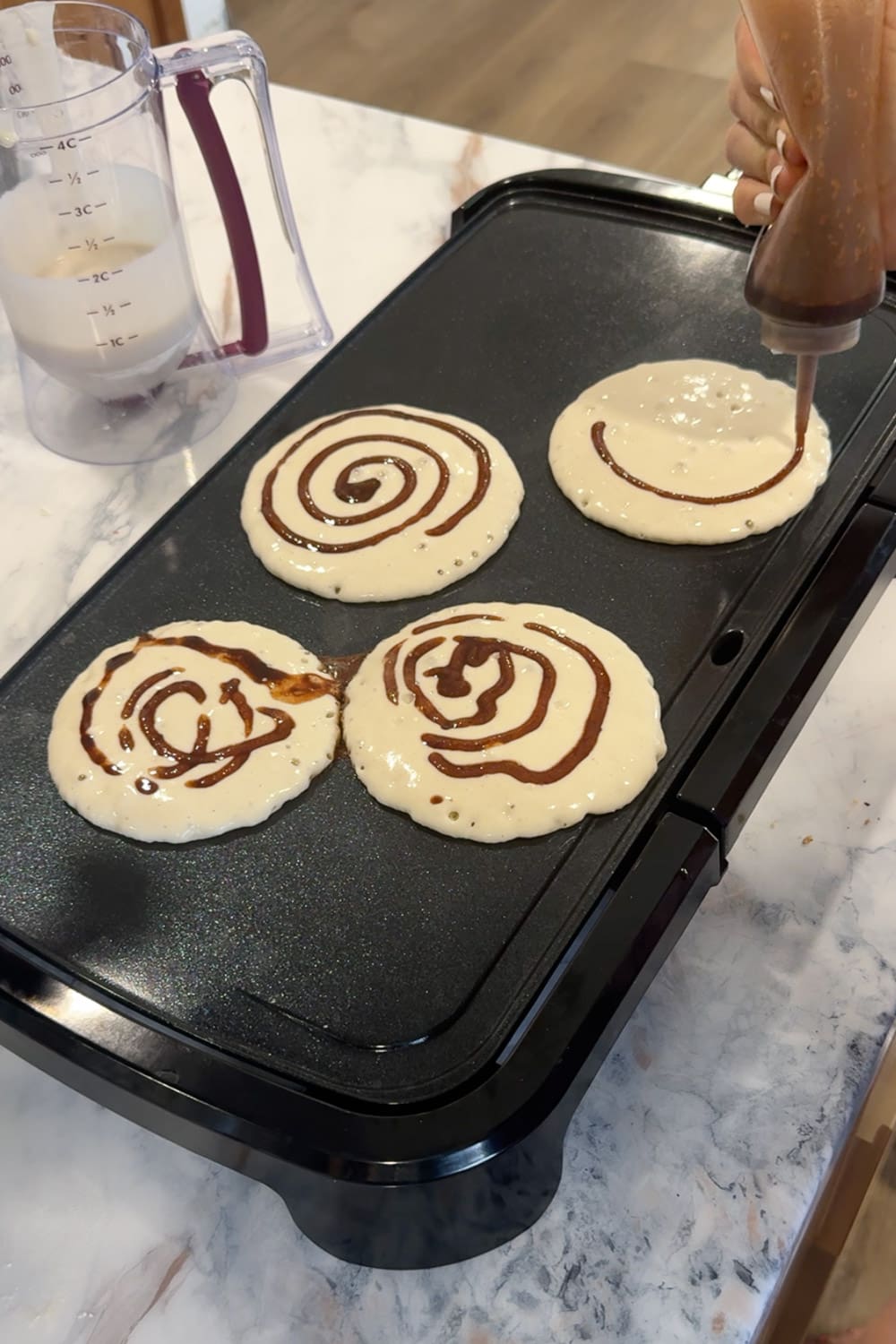 Hand using a squeeze bottle to pipe a cinnamon brown sugar spiral swirl onto four pancakes cooking on a black electric griddle, showing how to make the cinnamon roll pattern on cinnamon roll pancakes.