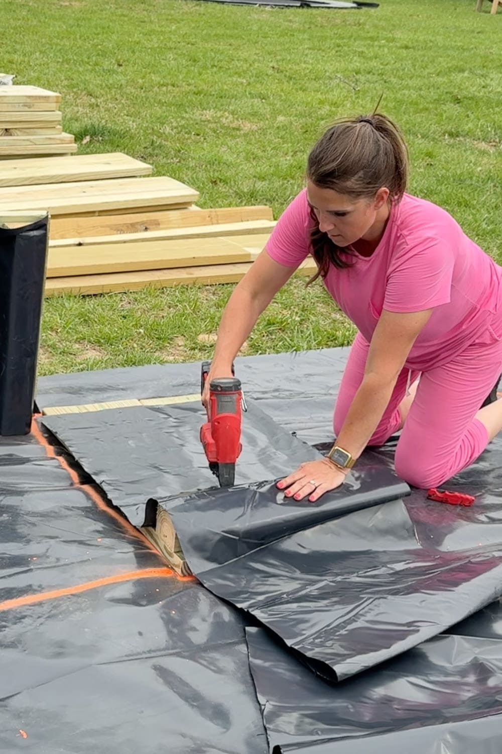A woman in a pink outfit kneels on black weed barrier fabric and uses a red nail gun to staple the liner flat to the base of a raised garden bed frame, with stacked pressure-treated lumber and orange spray paint layout lines visible nearby.