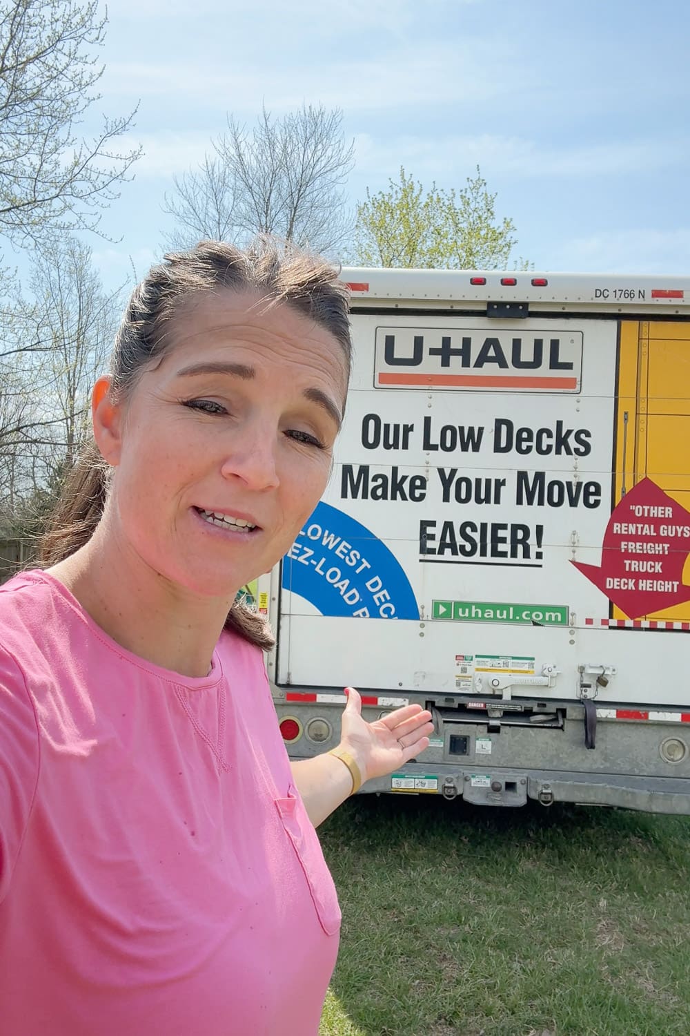A woman in a pink t-shirt gestures toward the back of a U-Haul rental truck parked on a grassy lawn on a sunny spring day, used to haul pressure-treated lumber for a planter box project.