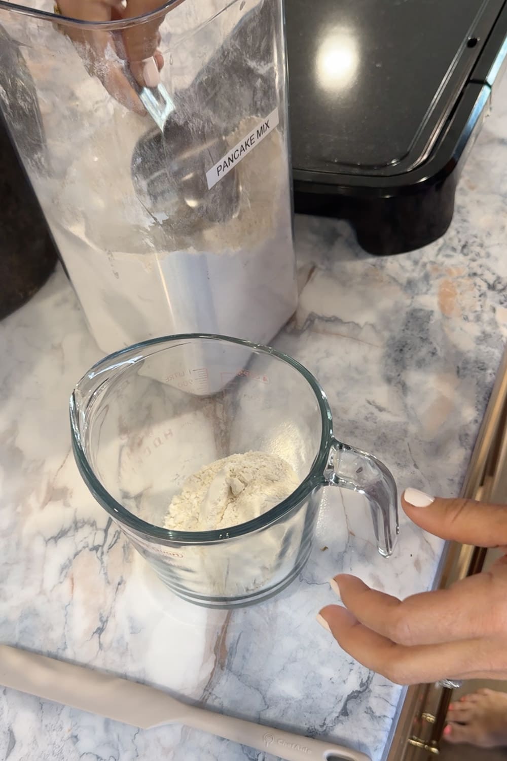 Hands scooping pancake mix from a labeled storage container into a glass measuring cup on a marble countertop, showing how to measure the pancake mix base for cinnamon roll pancakes.