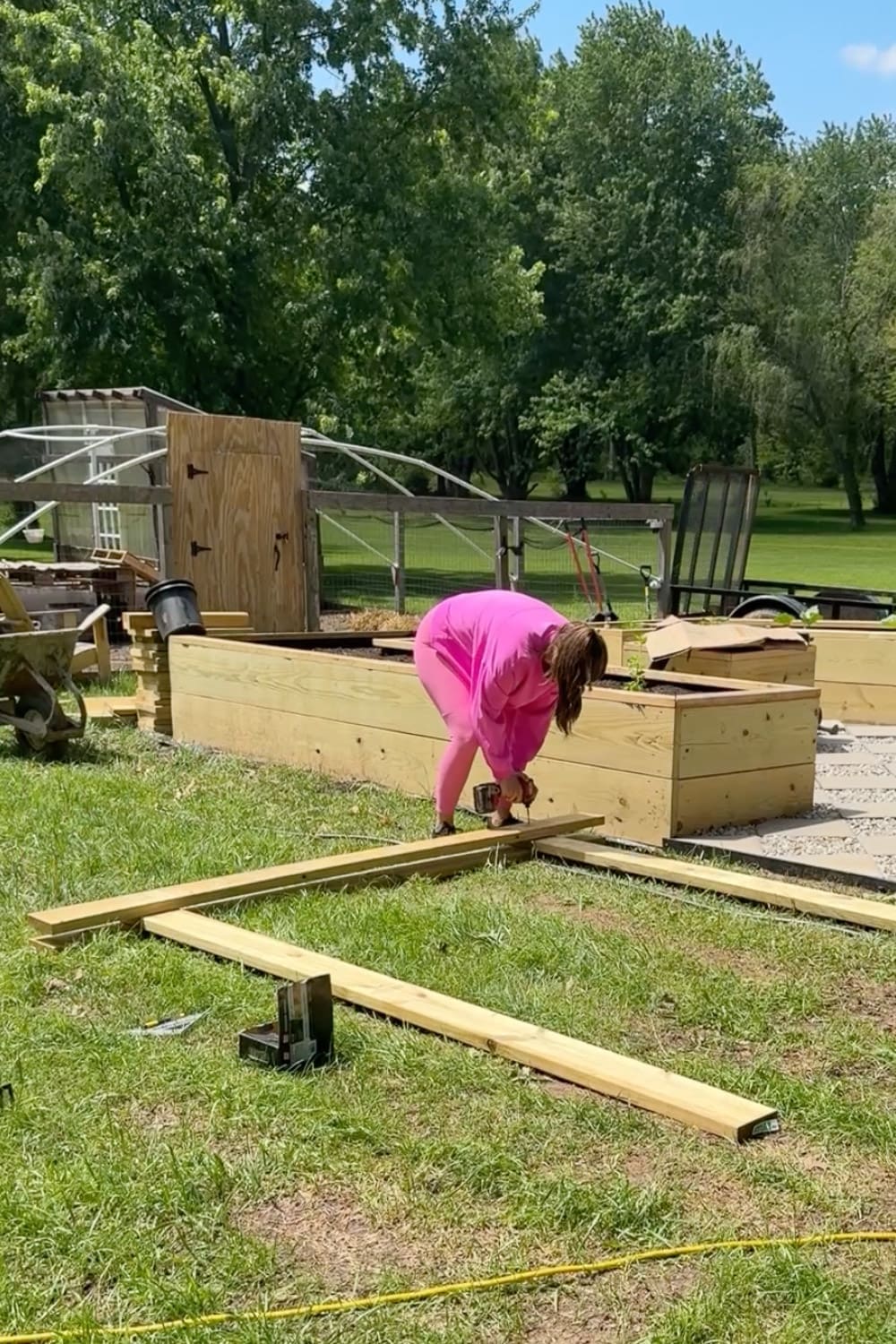 Woman in pink bending over to drill lumber pieces together while building a wooden garden arbor frame in the backyard near raised garden beds and a chicken coop