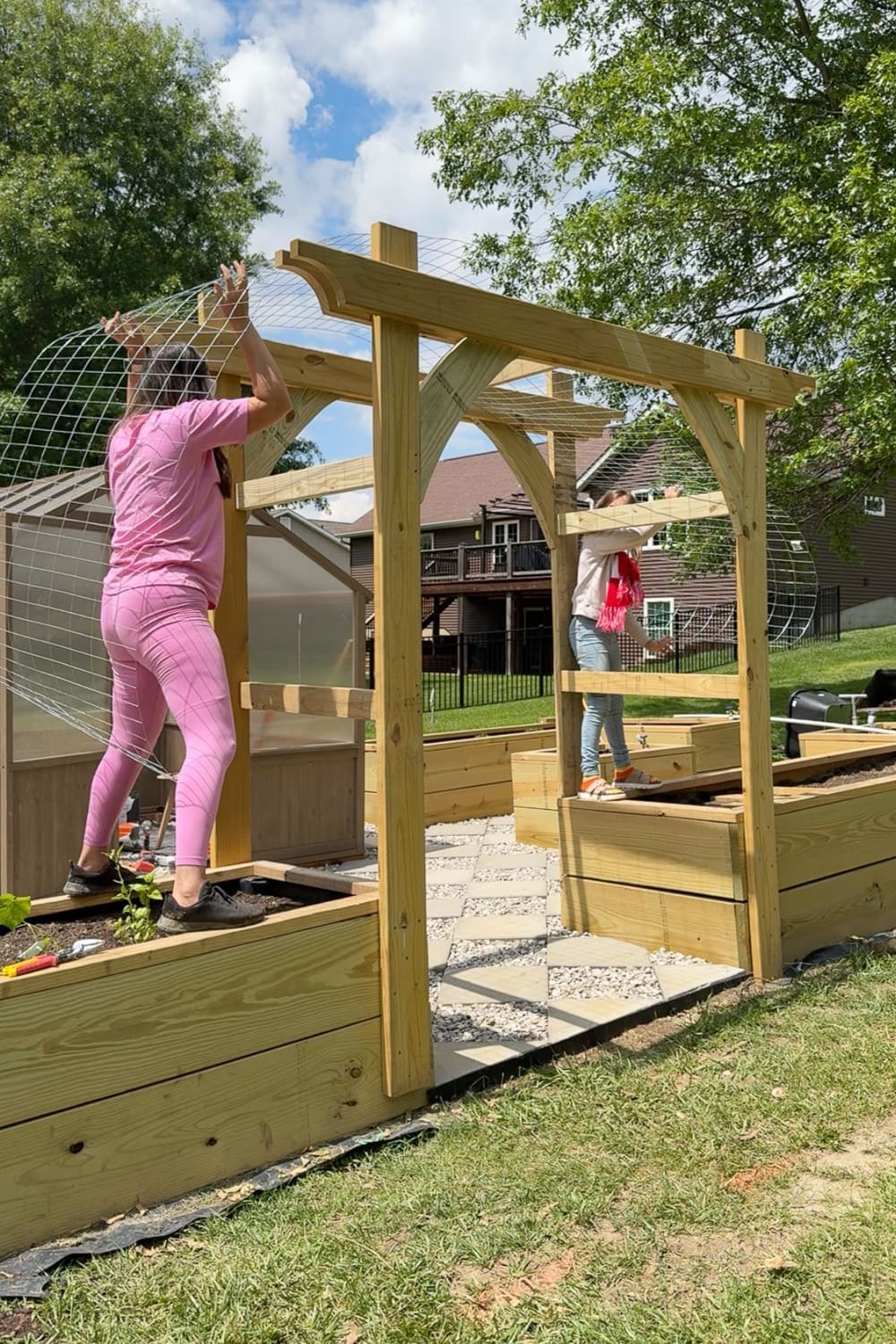 Two people working together to attach wire mesh trellis panels to a wooden DIY garden trellis arbor for climbing plants, standing on raised garden beds on either side of the arbor entrance