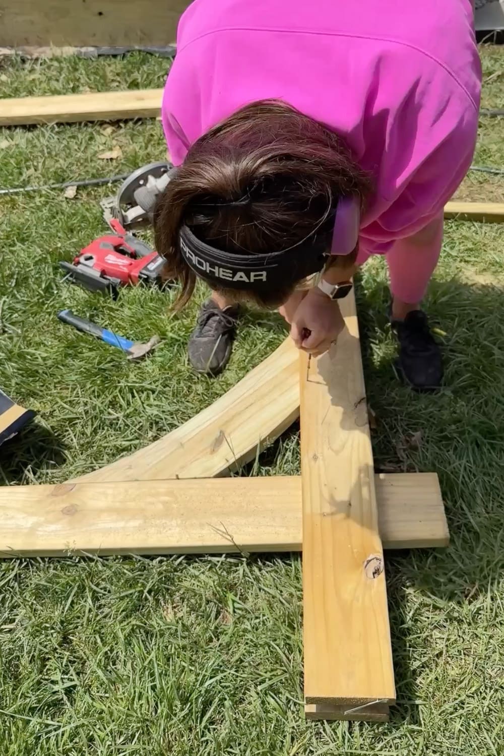 Woman in pink wearing ear protection assembling the wooden corbel and post pieces of a garden arbor on the grass with a circular saw and hammer beside her
