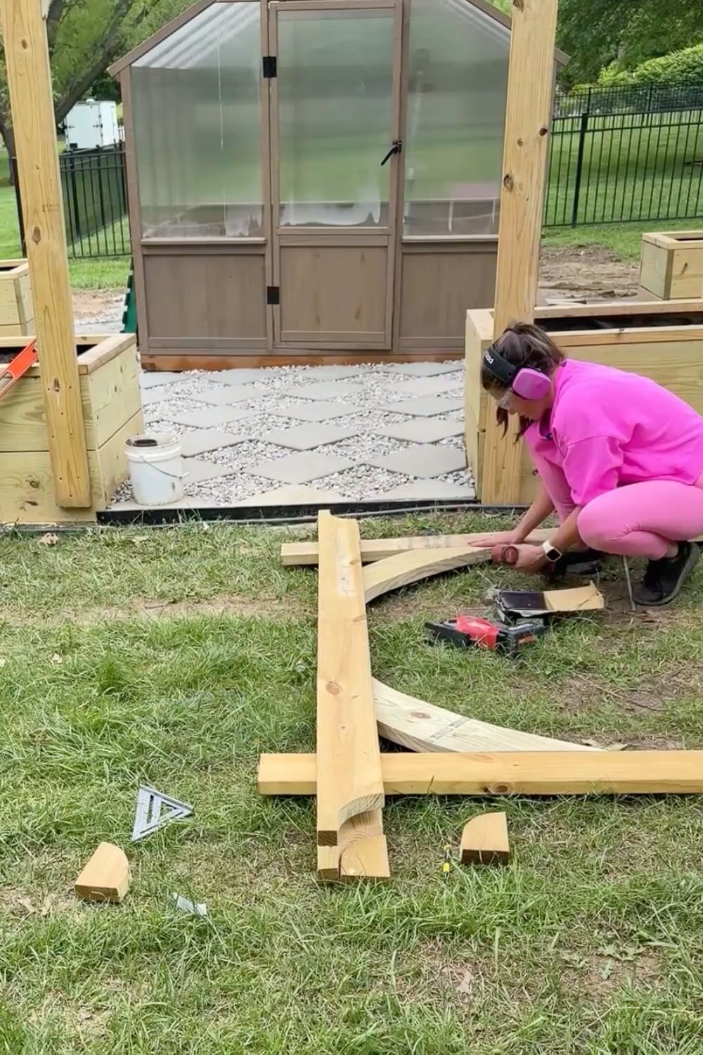 Woman in pink kneeling on the grass assembling the wooden frame and curved corbels of a DIY garden trellis arbor with the greenhouse and partially built arbor posts in the background
