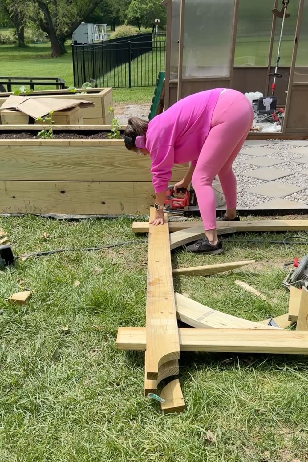 Woman in pink wearing ear protection using a cordless jigsaw to cut decorative curved ends into the long horizontal top beam of a DIY garden trellis arbor laid out on the grass