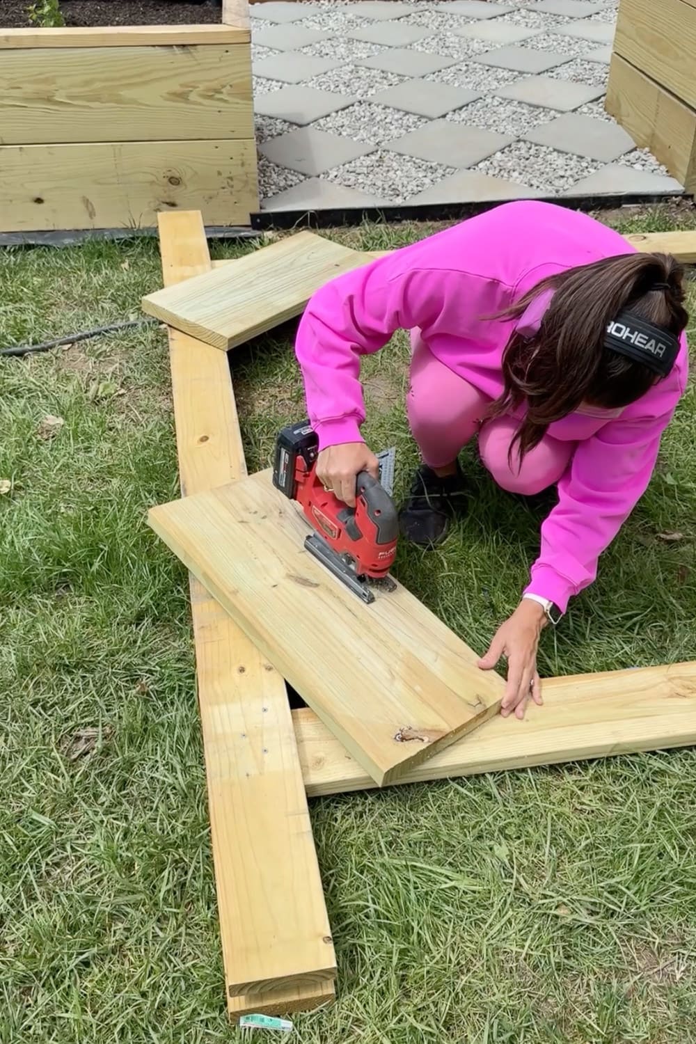 Woman in pink wearing ear protection using a jigsaw to cut a decorative curve into a wooden board for a DIY garden trellis arbor, kneeling on the grass next to raised garden beds