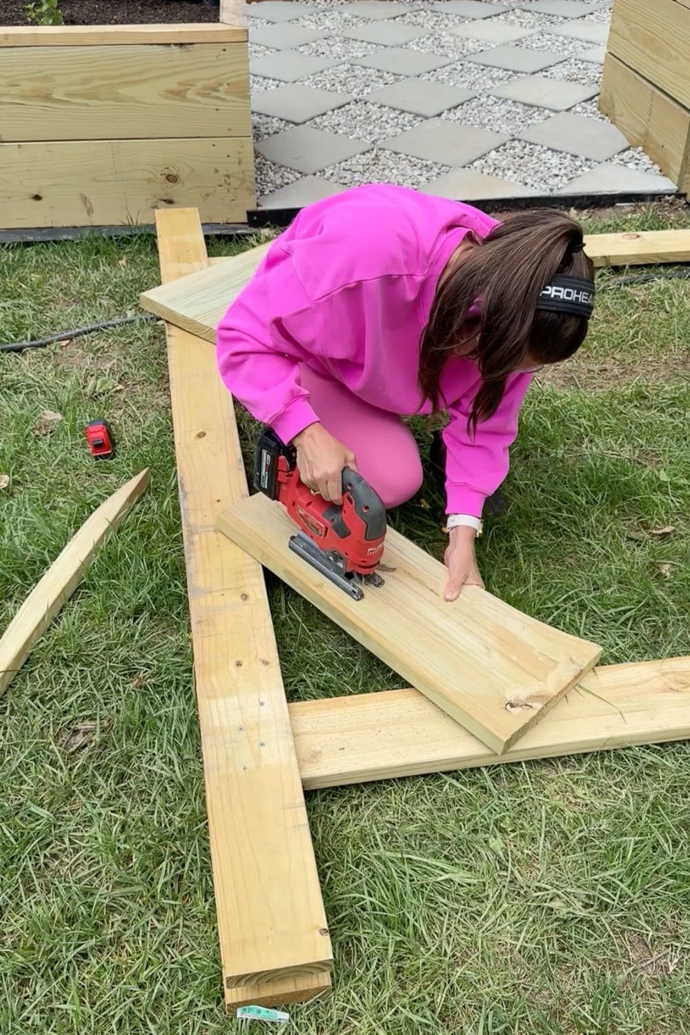 Woman in pink using a cordless jigsaw to cut along the marked curved line on a wooden board for a DIY garden trellis arbor corbel, with raised garden beds and a paver pathway in the background
