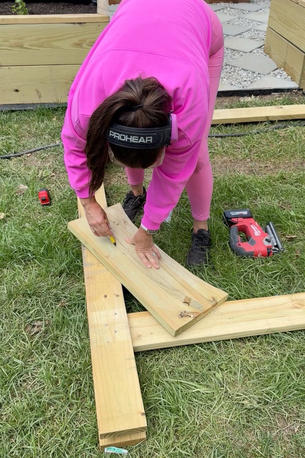 Woman in pink wearing ear protection drawing a curved line on a wooden board with a pencil to mark the decorative cut for a DIY garden trellis arbor, with a jigsaw nearby on the grass