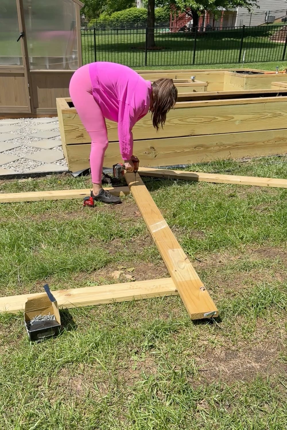 Woman in pink using a cordless drill to assemble the wooden frame of a DIY garden trellis arbor on the grass next to raised garden beds, with a box of screws nearby