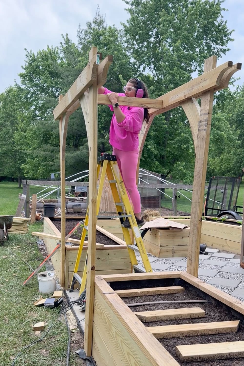 Woman in pink wearing safety glasses and ear protection standing on a yellow ladder to install the top crossbeam of a DIY garden trellis arbor between two raised garden beds