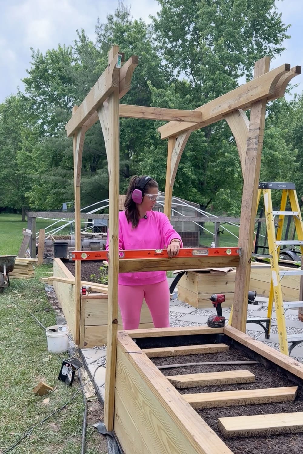 Woman in pink using an orange level to check that a horizontal support beam on the wooden garden arbor is even, with the assembled arbor frame standing over the raised garden beds
