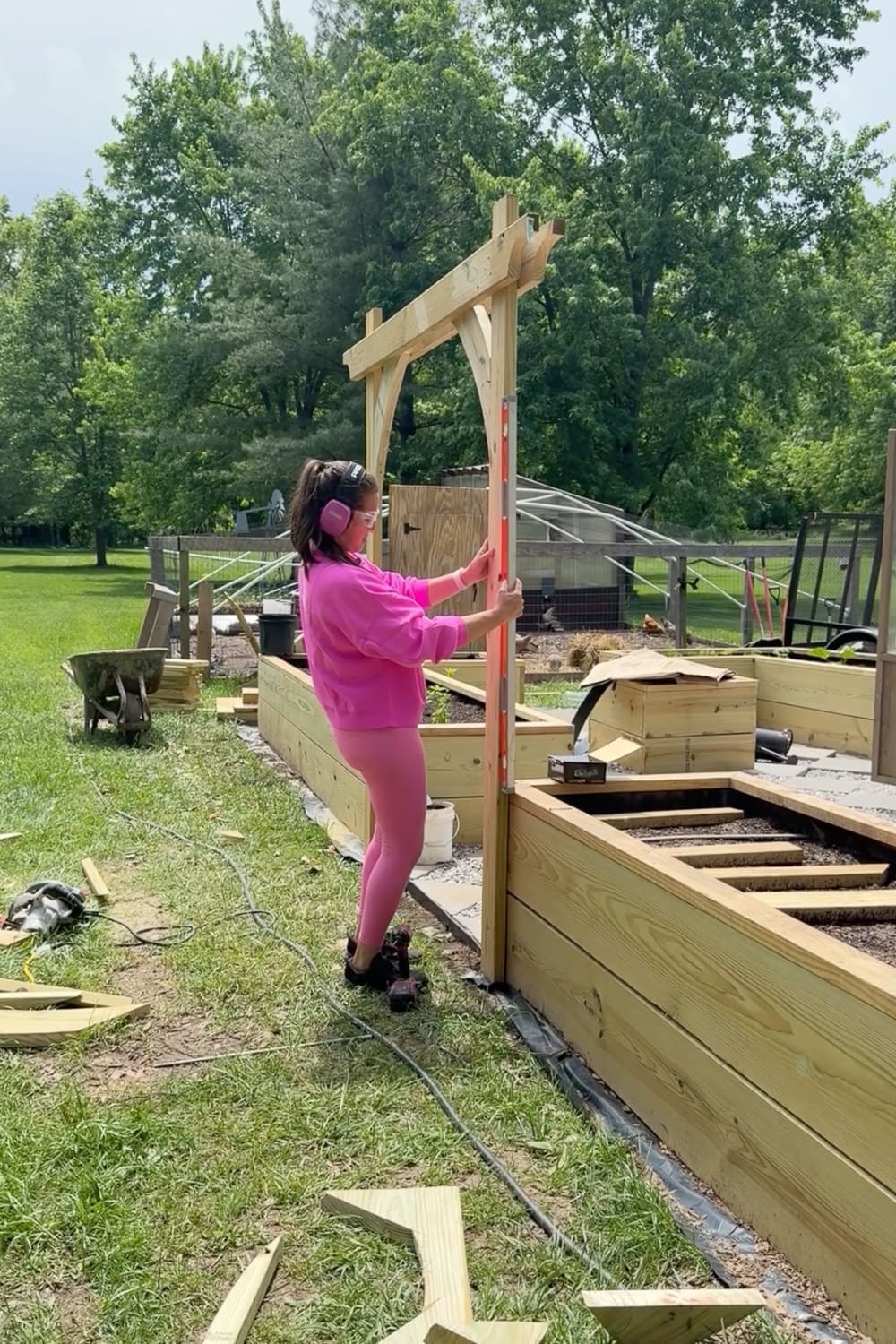 Woman in pink using an orange level to check that the upright post of a freshly assembled wooden garden arbor is plumb between two raised garden beds