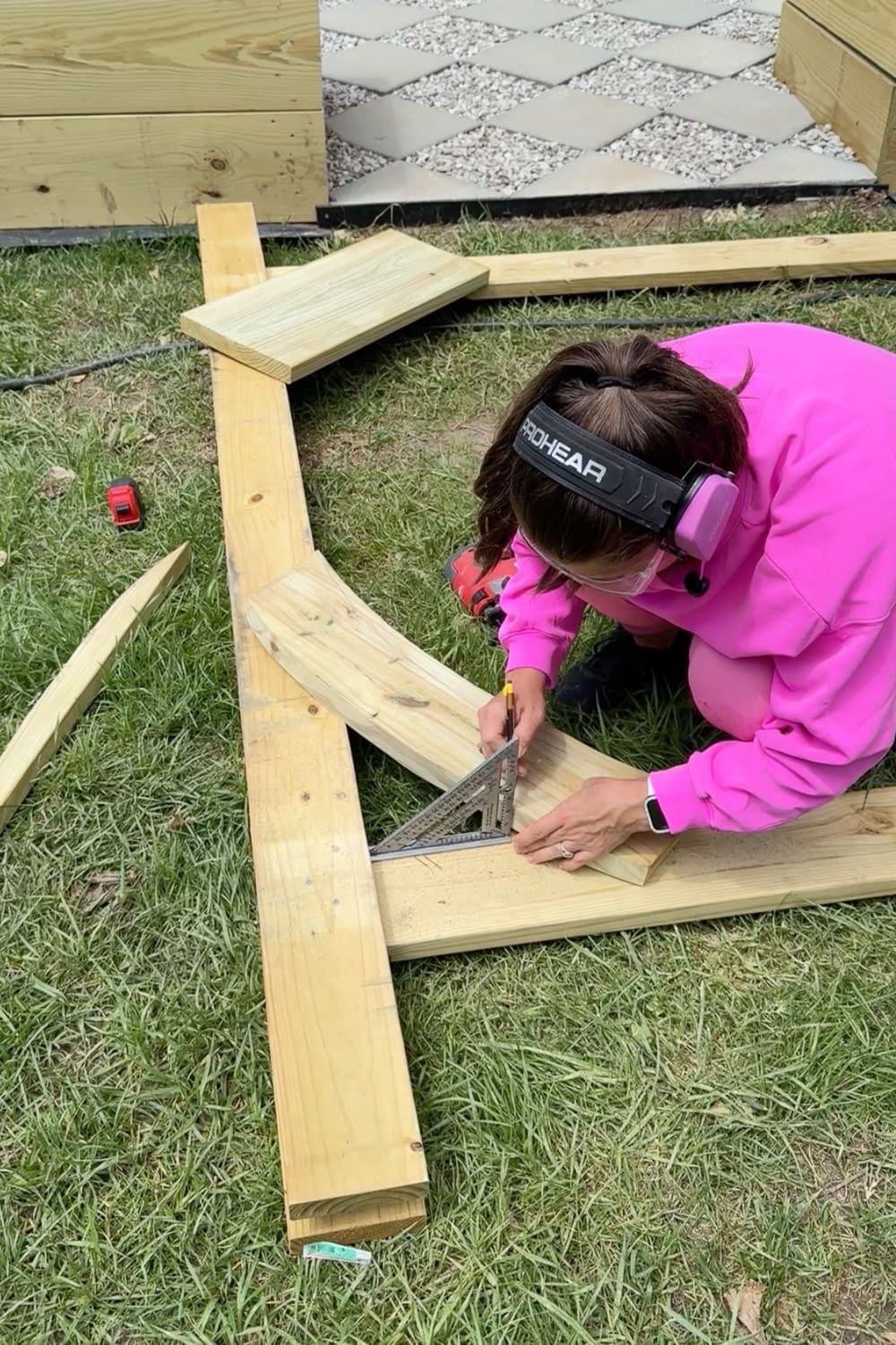 Woman in pink wearing safety glasses and ear protection using a speed square and pencil to mark angled cuts on a wooden corbel for a DIY garden trellis arbor