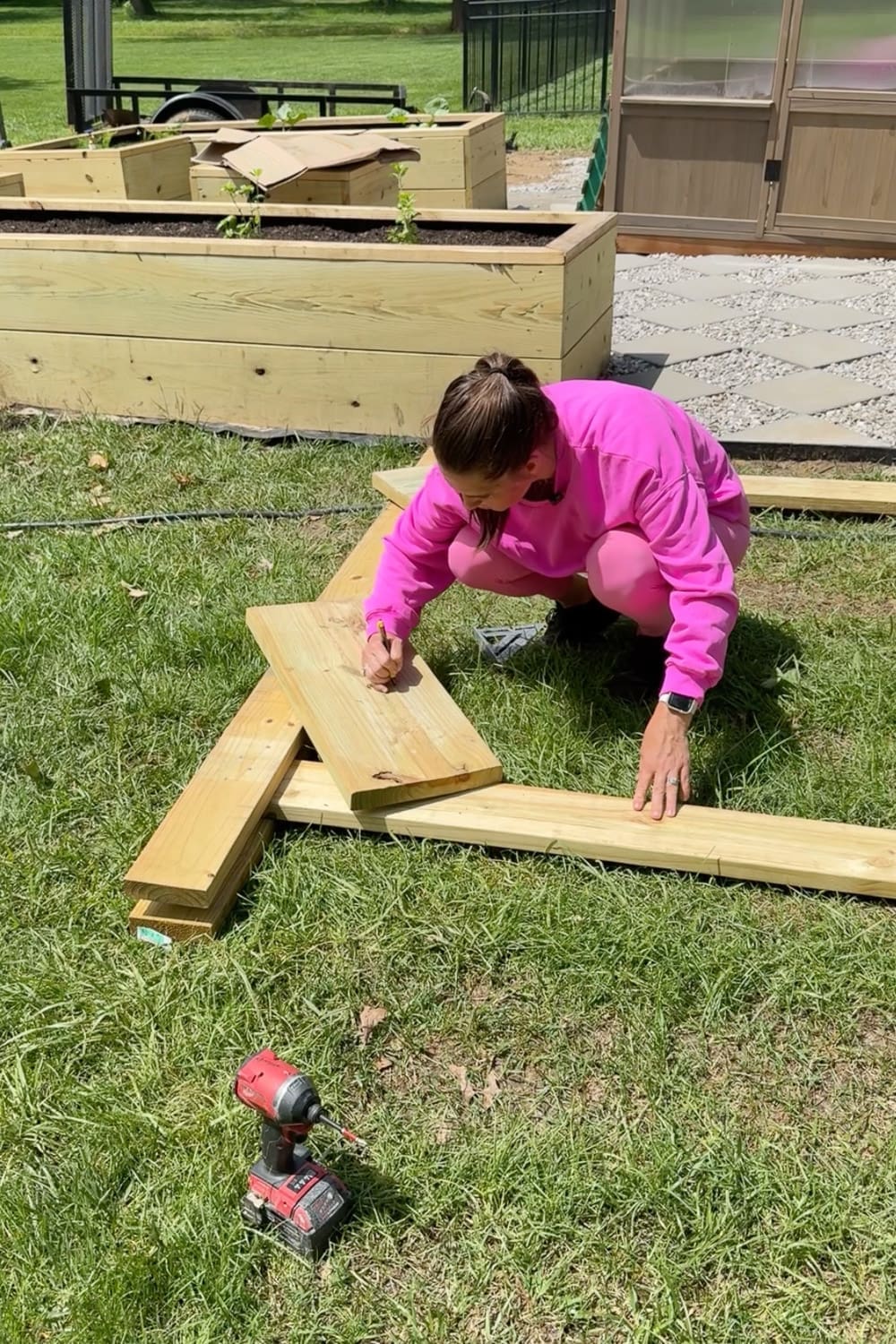 Woman in pink tracing a curved template onto a wooden board with a pencil to mark the decorative arbor corbel before cutting, with a cordless drill on the grass beside her