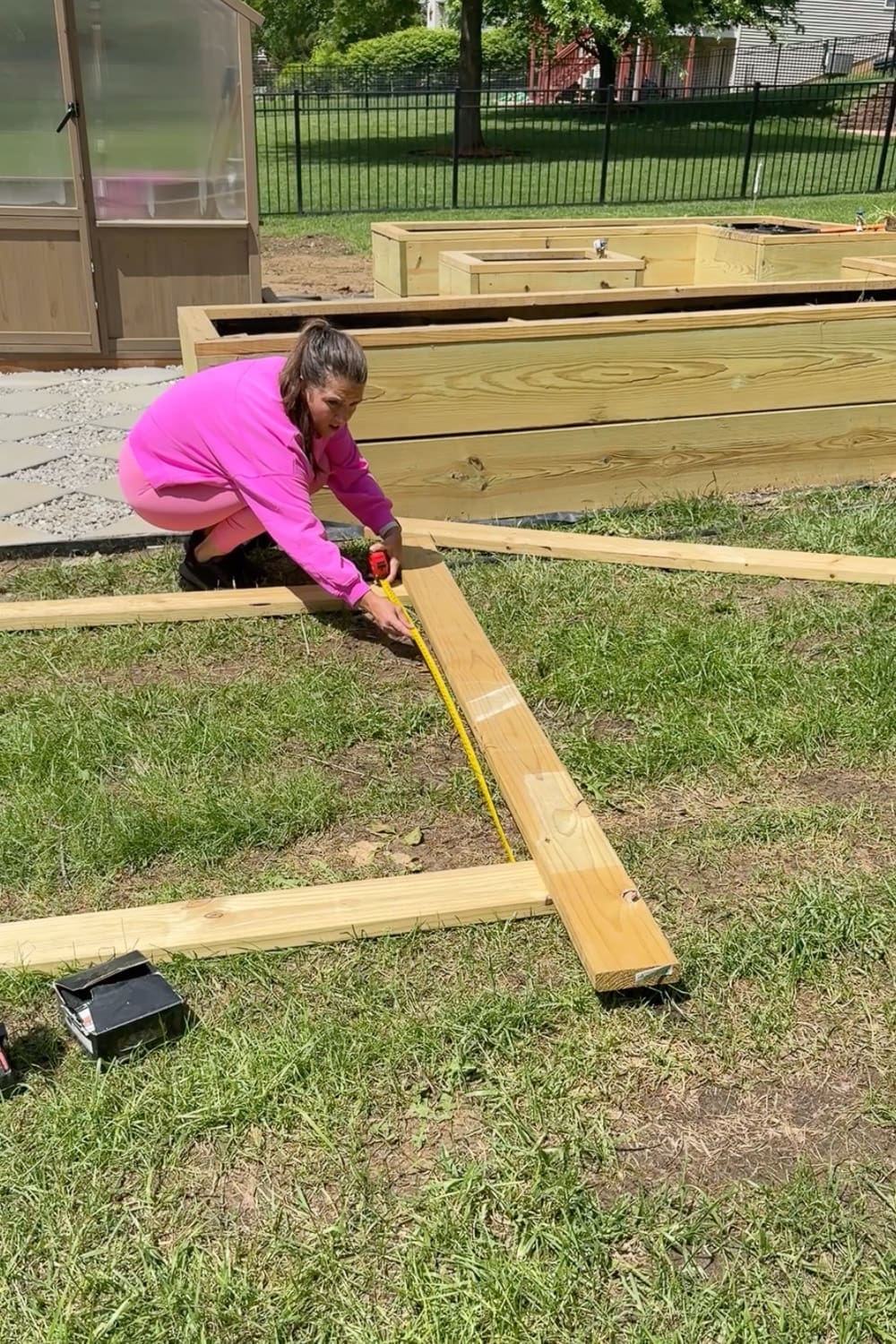Woman measuring lumber on the grass with a tape measure while planning the layout for a wooden arbor next to raised beds and a greenhouse