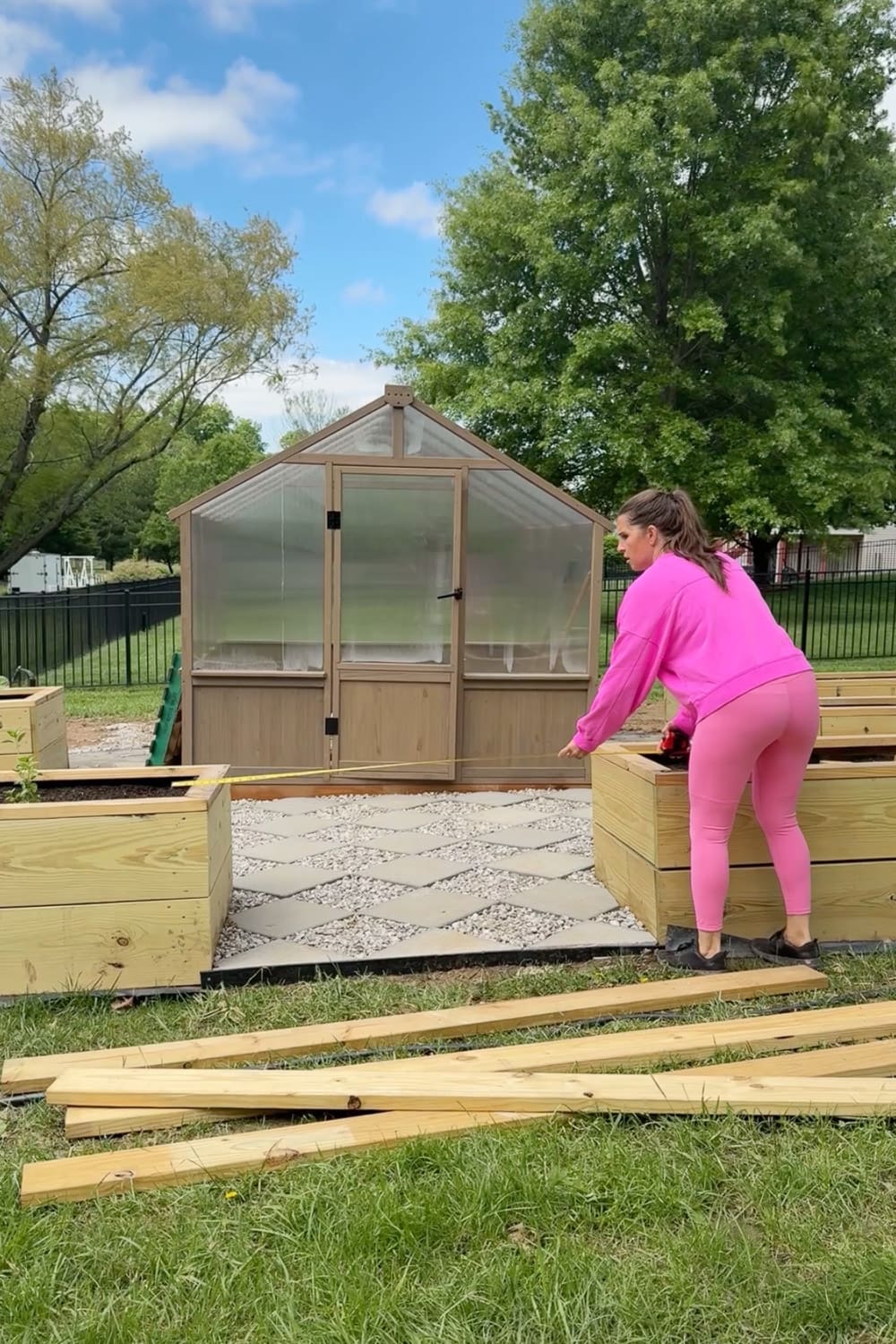 Woman in pink outfit measuring the space between raised garden beds with a tape measure to plan a DIY garden trellis arbor build, with greenhouse and lumber in the background