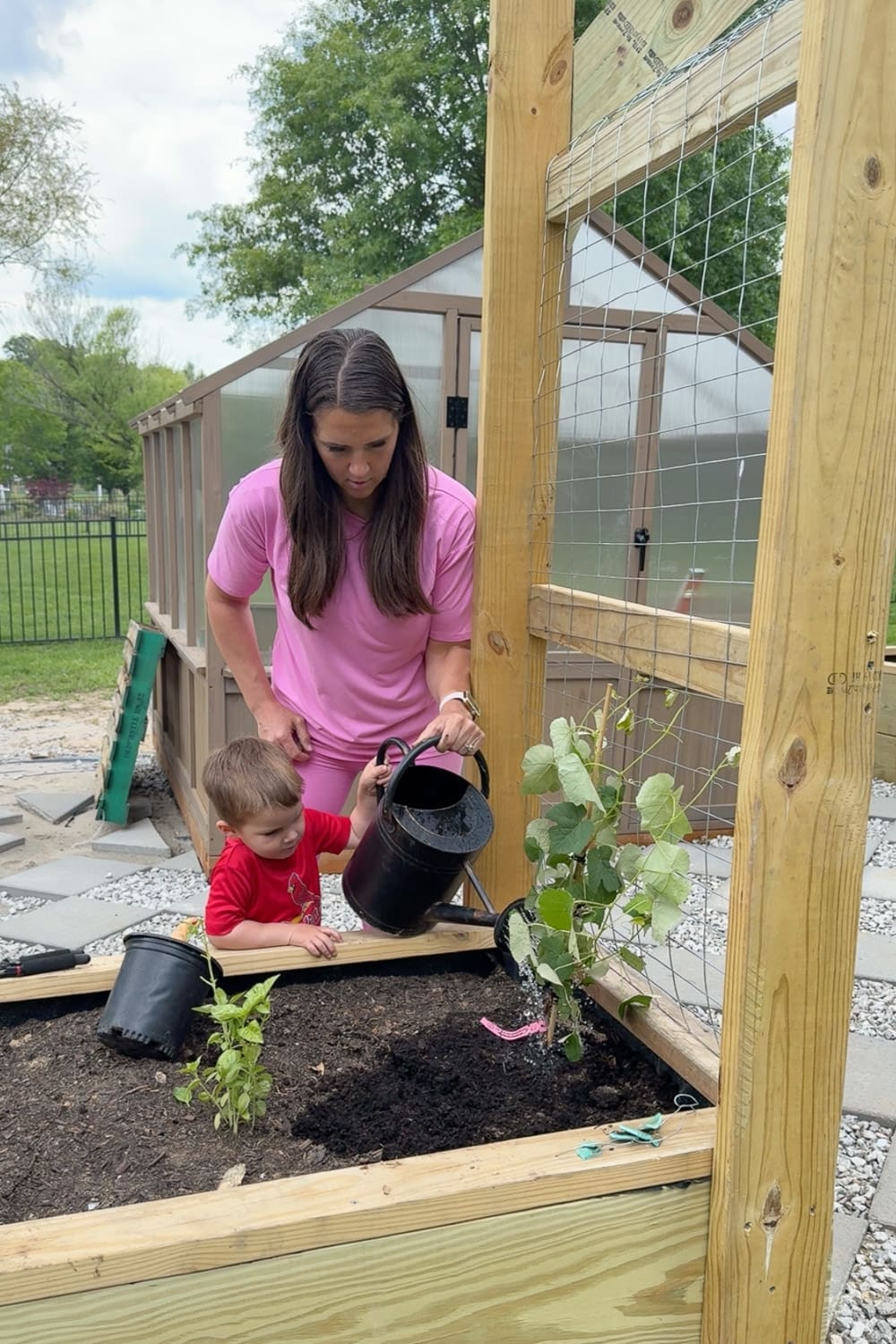 Mom in pink and young son in red shirt watering a newly planted climbing vine in a raised garden bed at the base of a finished DIY garden trellis arbor with wire mesh panels