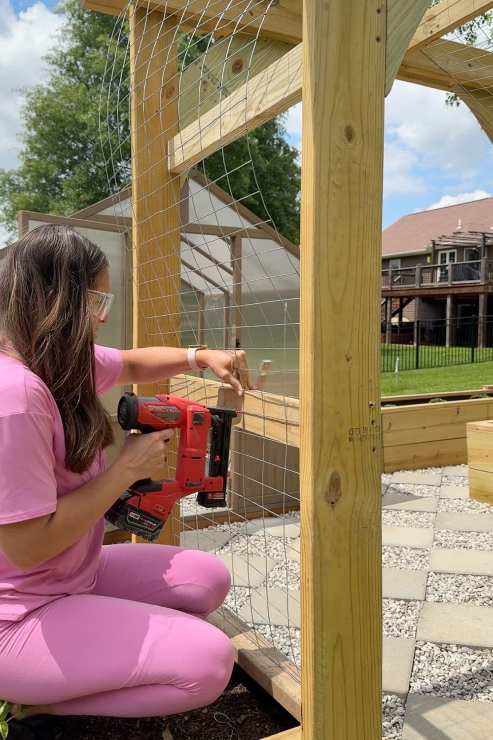 Woman in pink wearing safety glasses using a cordless crown stapler to secure wire mesh trellis panels to the wooden post of a garden arbor for climbing vines