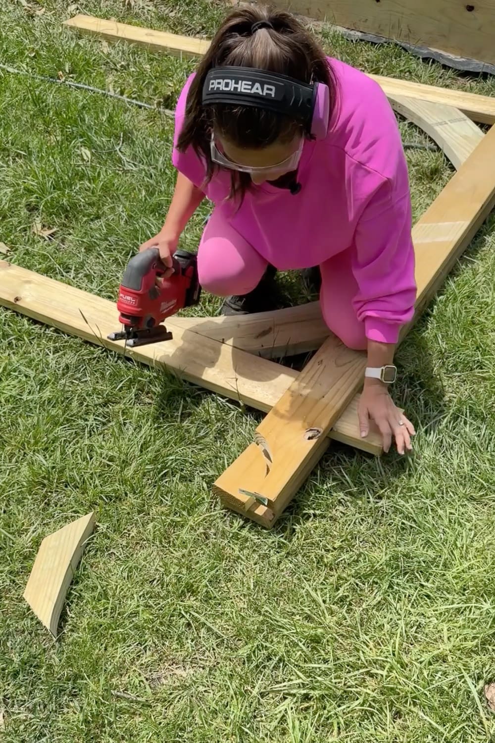 Woman in pink wearing safety glasses and ear protection using a cordless jigsaw to trim the end of a wooden board for a DIY garden trellis arbor frame